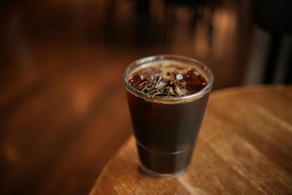Close-up of a cold brew coffee with ice cubes in a plastic cup on a wooden table indoors.