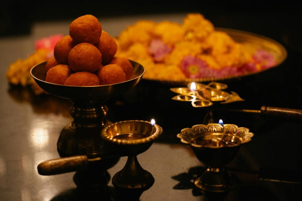 Close-up of Indian sweets with brass lamps and flowers on a dark background.