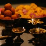Close-up of Indian sweets with brass lamps and flowers on a dark background.